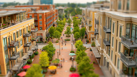 A tilt-shift view down a tree-lined main street with residential balconies, sidewalk cafes, umbrellas, and people walking the central pedestrian spine.